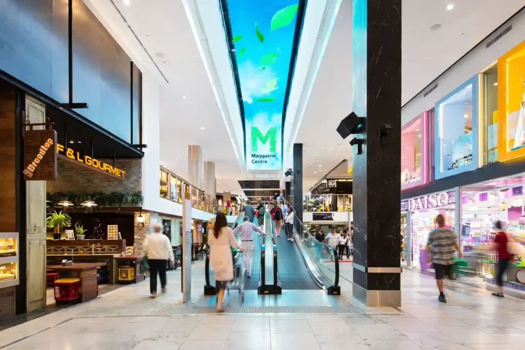 نمای داخلی مرکز خرید Macquarie Centre با مسیر متحرک، فروشگاه‌های دو طرف، سقف نورپردازی‌شده مدرن و حضور خریداران.
